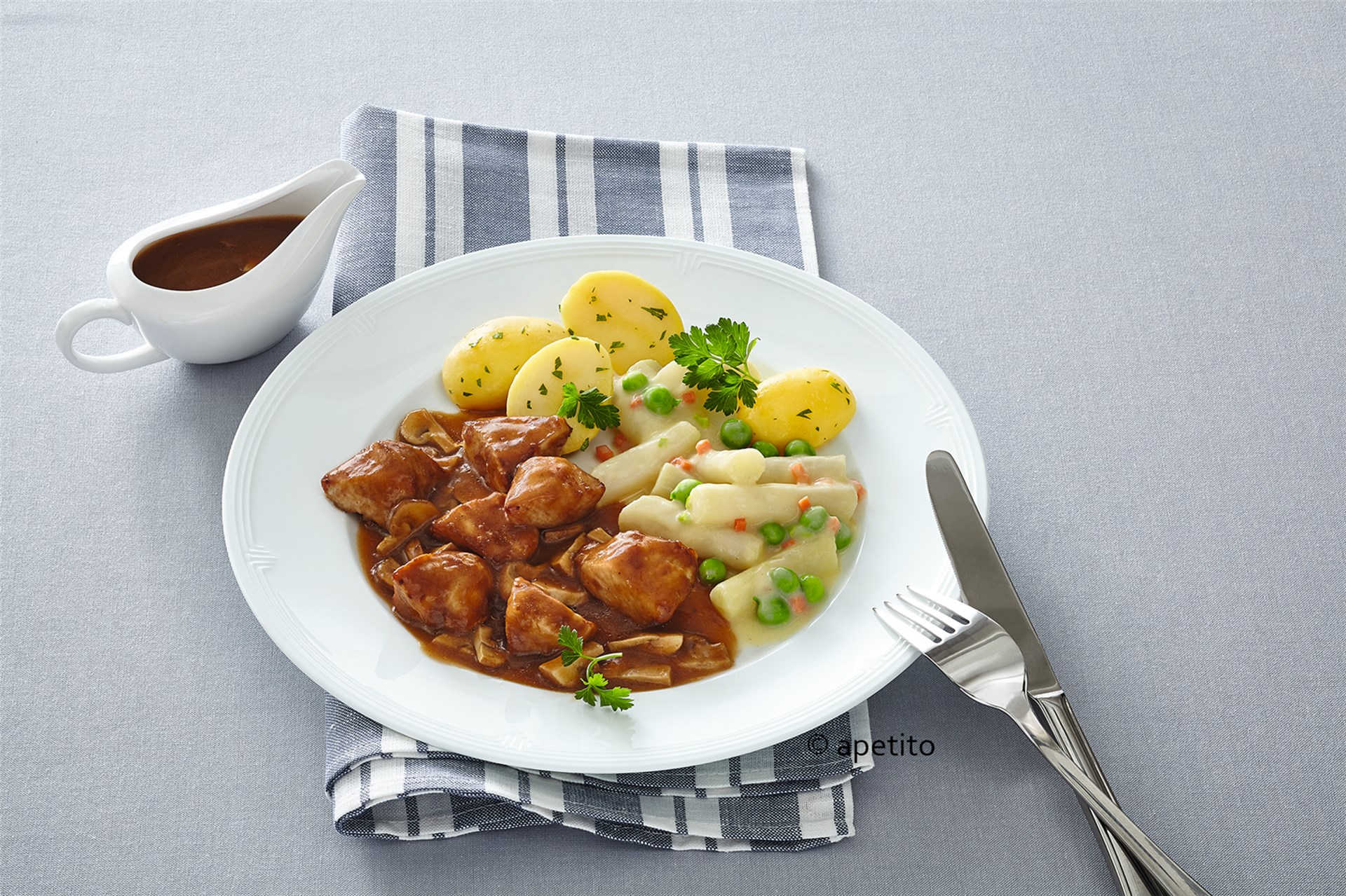 Das Bild zeigt einen angerichteten Teller mit einem vollständigen Gericht: Fleischstücke in brauner Soße, Salzkartoffeln mit Petersilie und ein Gemüse-Mix aus Erbsen und Karotten in weißer Soße. Der Teller steht auf einer blau-weiß gestreiften Serviette, rechts daneben liegen Besteckteile. Links im Bild befindet sich eine kleine weiße Sauciere mit zusätzlicher brauner Soße. Am unteren Tellerrand ist das Wort „apetito“ zu sehen.
