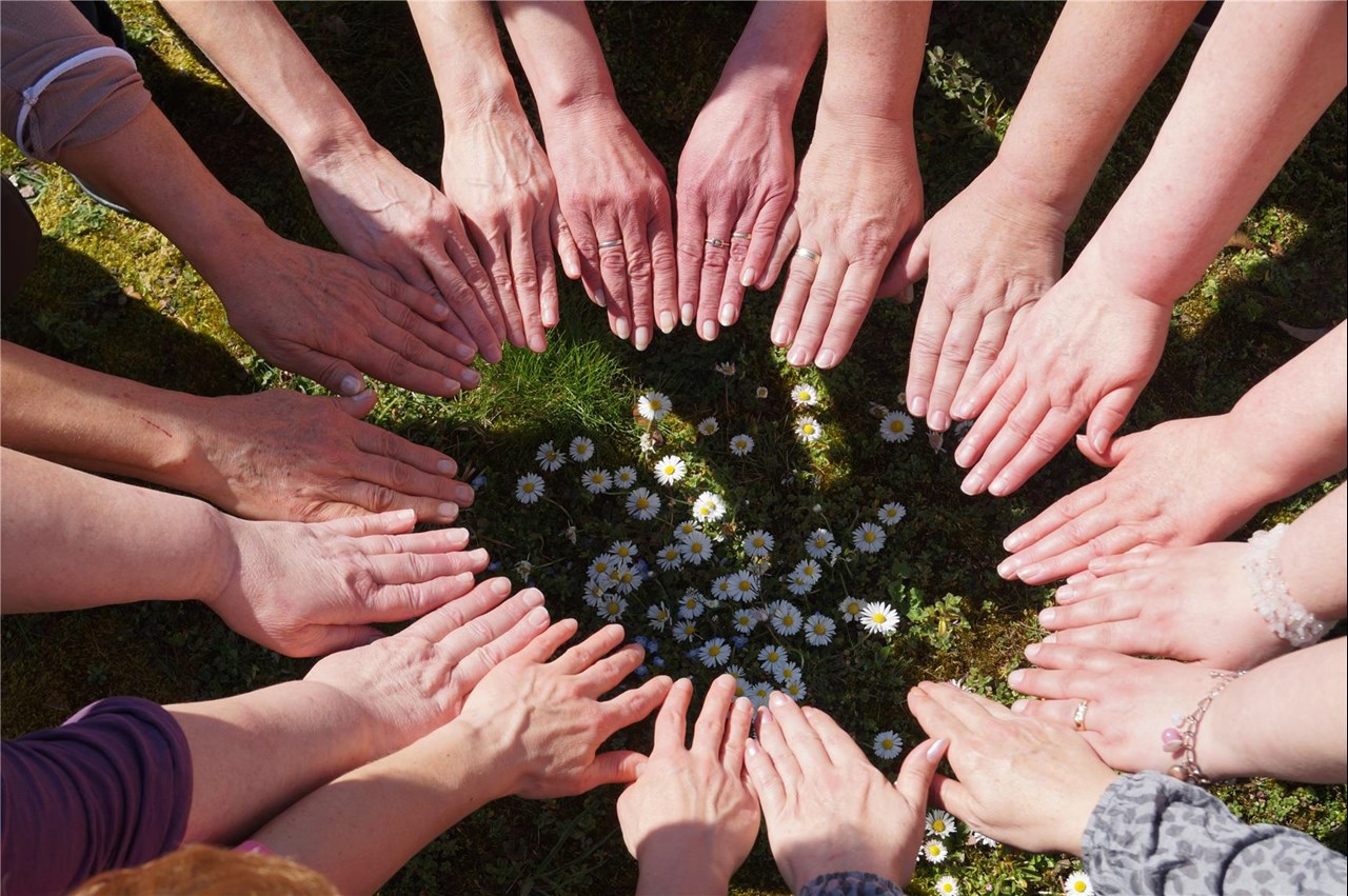 Das Bild zeigt eine Gruppe von Menschen, die im Kreis stehen und ihre Hände zur Mitte hin ausstrecken. In der Mitte liegen kleine weiße Blumen auf dem Gras.